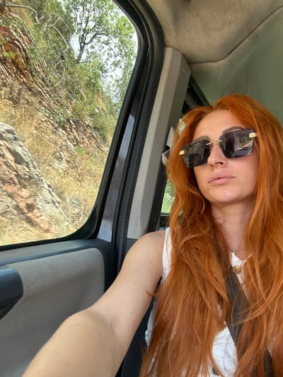 The image is a selfie taken inside a vehicle, oriented vertically. The foreground features a person with long, bright red hair, wearing large, square, dark sunglasses. Their head is tilted back, and their eyes are not visible behind the sunglasses. Their lips are slightly parted, and their skin appears fair. The person is wearing a white top with lace detailing. A delicate silver necklace with a small gold pendant is visible around their neck. Their arm is resting on the car's door frame.
The background shows the interior of the car, with a grey fabric-covered door panel. Through the open car window, a rocky, arid landscape with sparse green foliage and trees is visible. The sun appears to be bright, casting a warm light on the scene, suggesting it is daytime, likely late morning or afternoon. The overall impression is of a casual, outdoor excursion or road trip. The crops provide close-ups of the person's face and hair, as well as details of the car's interior and the landscape outside. There is no visible text in the image.