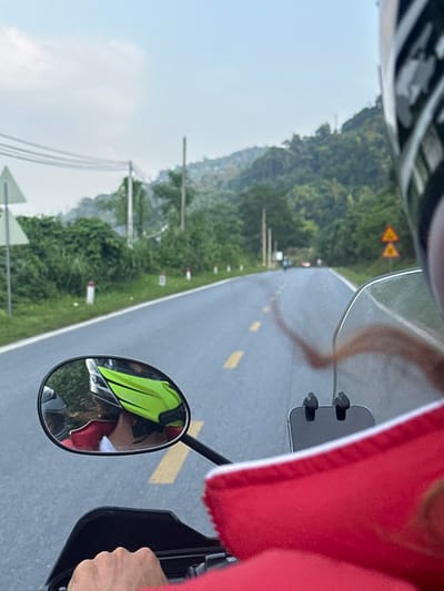 The image captures a first-person perspective from a motorcycle rider or passenger traversing a paved, winding road in a mountainous and verdant landscape, likely in Vị Xuyên Commune, Vietnam.
In the foreground, elements of the motorcycle and rider are visible. The top-left corner shows a part of a helmet with a black and white pattern. On the right, a red garment, possibly a jacket or sun protection, is prominent, and a hand firmly grips the motorcycle's handle. A clear windshield reflects a blurry, elongated shape that appears to be legs. A rearview mirror on the right offers a reflection of the person's torso, clad in a bright neon yellow vest or jacket over a red layer, confirming the presence of at least one person on the bike.
The main subject is the road ahead, stretching into the distance. It is a well-maintained asphalt road with distinct yellow dashed lines marking the center. The road curves gently through a landscape dominated by lush green mountains and hills covered in dense vegetation under a bright, clear sky. Utility poles with multiple power lines run parallel to the road on the left side, and white and red roadside markers are intermittently visible. In the mid-distance on the left, two yellow triangular warning signs with red borders are posted, featuring black symbols that indicate upcoming road hazards, likely curves or winding road sections.
The scene suggests an outdoor journey or road trip, likely during daylight hours given the bright illumination and clear visibility. The weather appears fair and sunny. The winding road and mountainous terrain align with the known characteristics of Vị Xuyên Commune, offering scenic views. No specific emotions are discernible, but the setting conveys a sense of travel and exploration.