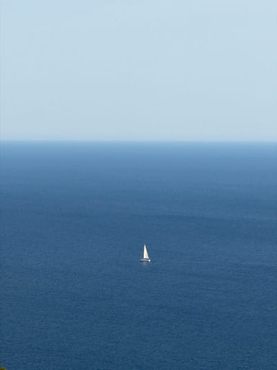 The image shows a vast expanse of deep blue sea under a clear, pale blue sky. The horizon line is clearly defined, separating the ocean from the sky. In the middle distance, a small white sailboat with a single sail is visible, appearing as a solitary object on the calm water. The scene is minimalist, emphasizing the scale of the ocean and the smallness of the boat within it. The location is identified as the sea off the coast of Krimovice, Montenegro.