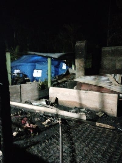 The image captures a night scene in Lhueng Tengoh, Indonesia, depicting the aftermath of significant damage, likely a fire. The foreground consists of dark, charred ground extensively covered with debris, including a distinct metal grate pattern, fragmented wooden planks, and various unrecognizable burnt materials.
In the midground to the right, the structural remnants of a building are visible. These include a prominent, dark, rectangular masonry column standing vertically, and a lighter-colored, rectangular base structure. Sections of corrugated metal sheeting, consistent with roofing material, are partially collapsed and rest against these remnants.
To the midground left, a large, blue tarp structure, resembling a temporary shelter or tent, is illuminated from within, casting bright rectangular glows through openings. This blue tarp is supported by light-colored, possibly green, vertical poles. The background beyond the structures is dark and indistinct, suggesting dense vegetation. No individuals are present in the scene.