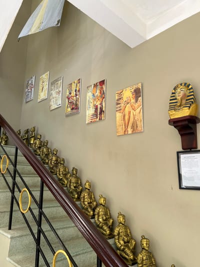 An interior shot features a stairwell decorated with historical art. Depictions of ancient Egypt are present, including a pharaoh bust and several rectangular images showcasing hieroglyphics and scenes from Egyptian life. A line of small, golden terracotta army statues flanks the staircase's railing. The cream-colored wall provides the backdrop for these cultural artifacts. A framed document hangs near the pharaoh bust.