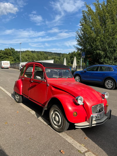A bright red, vintage Citroën 2CV is parked on the side of a street in Heilbronn, Germany. The car is facing towards the viewer's left, with its front slightly angled towards the camera. The car has a black convertible top. To the right of the Citroën, a modern blue SUV is parked further down the street. Behind the cars, there is a building with large windows and a fence. Further in the background, a treeline with lush green foliage is visible against a bright blue sky with scattered white clouds. A camping trailer is parked on the left side of the street, further in the distance. The street appears to be a paved road with a white line marking the edge of the parking area. The overall scene suggests a sunny day in a semi-urban or suburban environment. The license plate of the Citroën reads "HN TE 30H".