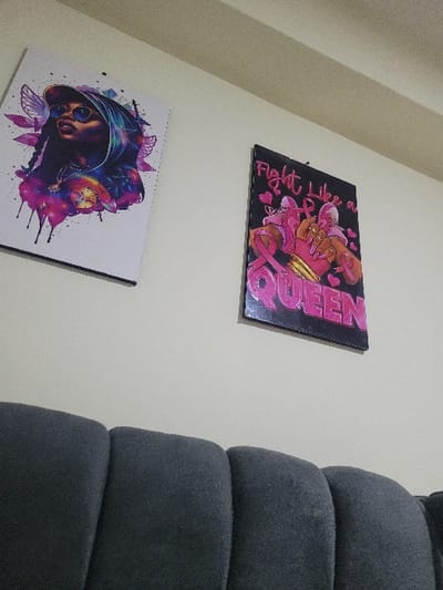 This image depicts an indoor scene in Nanyuki, Kenya, showing a section of a light-colored wall with two framed artworks mounted above a dark grey sofa. The upper portion of the sofa, specifically three distinct back cushions, is visible at the bottom of the frame. A ceiling beam is partially visible along the top edge of the image. The artwork on the left is a vertical rectangular poster featuring a stylized illustration of a person, likely female, with dark skin, wearing a cap and sunglasses, looking upwards. This illustration is rendered in vibrant shades of purple, blue, and pink, incorporating floral elements, wing-like details, and a watercolor-like drip effect. The artwork on the right, smaller and also vertically oriented, is a poster with a black border. It displays the phrase "Fight Like a QUEEN" in prominent pink lettering against a black background, surrounded by pink hearts, ribbons, and elements resembling boxing gloves.