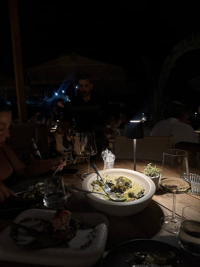 The image depicts an outdoor night dining scene at a restaurant in Heraklion, Greece. In the foreground, a wooden table is set with multiple items. On the left, a diner, partially visible, is actively eating with a fork and knife from a plate containing food remnants, including a piece of what appears to be lobster tail on a dark base. A large white bowl, centrally located, holds a partially consumed dish with green sauce and dark elements, accompanied by a fork and spoon. Two stemmed glasses filled with white wine, a clear glass of water, and a dark beverage bottle are visible. A black, modern-style portable table lamp illuminates the immediate dining area, flanked by silver-topped salt and pepper grinders and a small potted succulent plant. A mobile phone rests on the table near the foreground diner.

In the midground, a male server with a beard, dressed in dark clothing, stands facing the camera, positioned behind the primary table. Beyond him, other patrons are seated at additional dimly lit tables under large, light-colored umbrellas made of natural materials. The background is dark, with distant artificial lights visible, consistent with an open-air, possibly seaside, dining establishment.