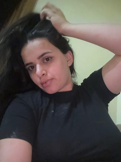 A casual, rotated portrait captures a person, likely a woman, with dark, long hair and a medium complexion, looking directly into the camera. She is positioned on her side or back, with her head resting on a surface. Her right arm is raised, with her hand lightly touching her hair near her head.

The subject wears a plain black short-sleeved top, which shows several small, light-colored specks, possibly dust or lint, on its surface. Her facial features include thick, dark eyebrows and dark brown eyes. Her lips are slightly parted, revealing a hint of her tongue, giving her expression a neutral to slightly playful or pensive quality.

The background is simple, featuring a solid light yellow-beige wall, with a darker wooden surface visible in the lower-left corner of the frame. The lighting appears soft and even, indicative of an indoor setting. The image lacks specific location cues, though it is contextualized as being taken in Ghukasavan, Armenia. No visible text is present in the media file. The overall impression is that of a candid or self-shot photograph.