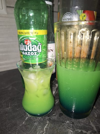 The image shows two bottles of Uludağ Gazoz, a Turkish carbonated soft drink, and a glass filled with the same green-colored beverage with ice. The bottle on the left is mostly full, while the bottle on the right appears to be empty or nearly empty. The drink in the glass is a vibrant green, and the bottle labels are predominantly green with white and yellow text. Visible text on the bottles includes "Uludağ GAZOZ SEIT 1930" and "ORIGINAL GAZOZ". The image is oriented vertically, suggesting it may have been taken with a mobile phone. The setting appears to be indoors, possibly a kitchen counter or a table, with a dark, textured surface in the background. The lighting is artificial, and there are reflections on the glass and bottles, indicating a possible indoor environment. The overall impression is one of refreshment and casual consumption of a beverage. There are no people visible in the image. The environment suggests a domestic setting in Bochum, Germany, given the context.