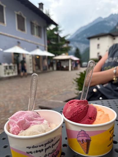 Two cups of gelato are presented in an outdoor setting, likely in Molveno, Italy, given the context. The cups are yellow and white, decorated with a cartoon image of an ice cream cone. The first cup contains two scoops of gelato, one deep red and the other a lighter pinkish-red. A clear plastic spoon, labeled "COMPOSTABLE," rests on top of the red scoop. The second cup, partially visible, holds scoops of pale pink and light cream-colored gelato, with a similar compostable spoon.

In the background, out of focus, a person wearing a dark t-shirt is visible, with a bracelet on their arm. Beyond the person and the cups, a blurred scene unfolds. A building with multiple windows, possibly a cafe or shop, is visible. Further in the distance, a large mountain looms under a cloudy sky, suggesting a cooler or overcast day. The overall impression is a casual moment enjoying gelato outdoors, perhaps on a vacation or outing. The visible text on the spoons confirms they are made of compostable material. The text on the cups is partially visible and appears to be in Italian, with phrases like "mi dai un gelats?" (Will you give me a gelato?) and "un cono per favore" (a cone please).
