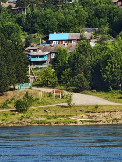 The image shows a cluster of wooden houses nestled into a forested hillside overlooking a wide, blue river. The houses are of varying sizes and architectural styles, with some featuring bright blue or gray roofs. A dirt road winds its way up the hill, passing by a small turquoise fence and a utility pole. Dense green trees surround the houses, with some reaching up towards the sky. In the foreground, the river is calm, with gentle ripples on its surface. The overall impression is one of a peaceful, rural setting. The scene is likely located in Pristan', Russia, a town known for its natural beauty and proximity to the Yenisei River.