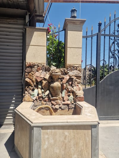 In Nor Kharberd, Armenia, a stone fountain is the central focus of the image. The fountain has a hexagonal base made of light-colored stone, with darker grey stone accents. Above the base, it transitions into a rock-and-stone wall adorned with a decorative urn and several colorful stones, including pink, brown, and green. Behind the fountain, a black metal gate with ornate scrollwork stands tall, topped with pointed finials. To the left of the fountain, a grey roll-up door is visible. A clear blue sky serves as the backdrop for the scene, with the bright sun casting shadows on the ground. A rusty pipe runs horizontally above the scene, supported by a metal structure. A small lamp fixture is mounted on top of a stone pillar to the right of the fountain.