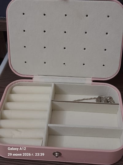 An open, pink jewelry box is displayed from a slightly elevated perspective. The interior of the box features several compartments. The top lid of the box is open and lined with a cream-colored, textured material that has numerous small, black, circular indentations. The bottom portion of the box contains two main sections. The left section is divided into multiple rows of cream-colored, padded ring holders. The right section is a larger, open compartment, currently empty except for a delicate silver chain with two connected charms lying across its bottom. The jewelry box is placed on a dark, textured surface. In the lower-left corner of the image, text indicating "Galaxy A12" and "29 июня 2026 г. 23:39" is visible. The location is the city of Kara-Köl, Kyrgyzstan.