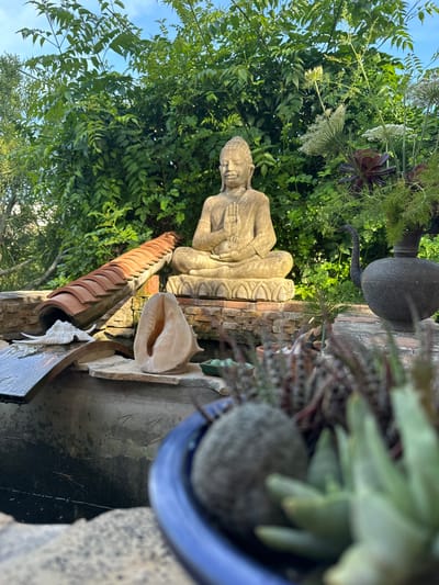 The image captures a serene outdoor scene in Altea, Spain. A weathered stone Buddha statue sits amidst lush greenery, suggesting a garden or tranquil courtyard. Beside the statue, a conch shell rests on a tiled surface. An aged ceramic pot is positioned nearby. In the background, there is dense foliage. A water feature is also visible. The overall composition evokes a sense of peace and natural beauty, consistent with Altea's Mediterranean atmosphere.