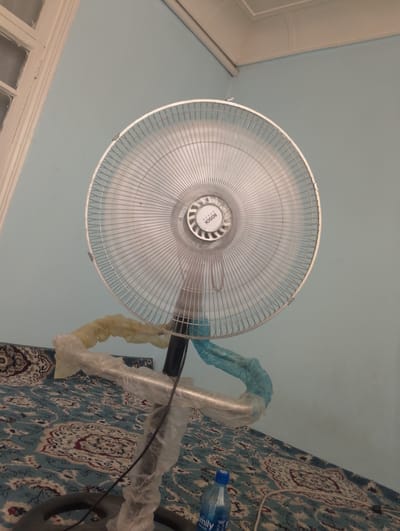 An indoor, eye-level shot captures a white Bosch brand pedestal fan positioned on a carpeted floor. The fan is predominantly white with a silver metal grille and a black stand. The stand and parts of the fan's base are wrapped in translucent plastic, with sections of yellow and blue plastic also visible around the stand. A blue plastic water bottle with a white cap sits on the carpet in the lower right corner of the frame. The wall behind the fan is painted a light blue. The image appears to be taken in a residential setting within Kokand City, Uzbekistan, based on the context provided.