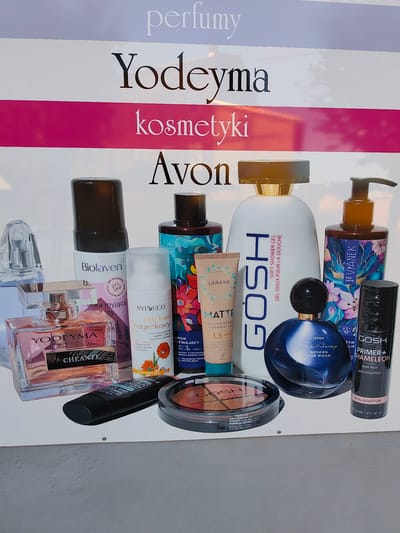 The image displays a promotional banner for beauty products, featuring various brands like Yodeyma, GOSH, Lumene, Sylveco, and Avon. The banner is situated in Stryków, Poland, specifically at the address 3 Maja 12. The products showcased include perfumes, lotions, shower gels, foundations, and makeup palettes. The banner itself is white with large text indicating "perfumy" and "kosmetyki," suggesting it is a display for a perfumery or cosmetics store. The products are arranged in a visually appealing manner, with a focus on bright colors and attractive packaging. There are no people visible in the image, and the environment appears to be a flat surface, possibly a wall or a display board. The lighting is even, suggesting an indoor or shaded outdoor setting.