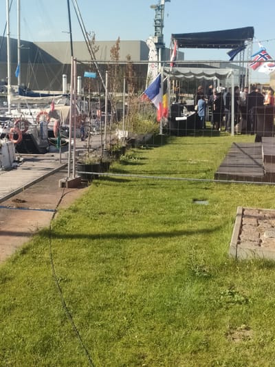 The image depicts a waterfront scene likely in Gdańsk, Poland, judging by the presence of flags from various nations, including Poland and possibly Belgium, France, Norway, and South Korea, suggesting an international event. In the foreground, a grassy area is cordoned off by a wire fence. Beyond the fence, several boats are docked, some with lifesavers visible on their decks. Further back, a covered stage with lighting equipment is set up, and a group of people are gathered underneath it. A large industrial building and a construction crane are visible in the background, indicating an active port or industrial area. The sky is clear and blue, suggesting a sunny day.