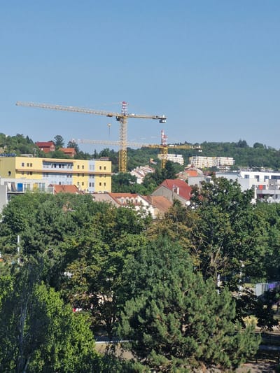 The image shows a cityscape with construction cranes against a clear blue sky. In the foreground, lush green trees dominate the lower half of the frame. Behind the trees, several buildings are visible, including a bright yellow building with multiple windows and balconies, and other residential and office buildings with various architectural styles and colors. Two large yellow construction cranes are prominently featured in the mid-ground, indicating ongoing construction activity. In the background, more buildings and rolling green hills suggest a suburban or semi-rural setting within a city. The overall scene depicts a developing urban environment with a mix of established structures and new construction, set against a backdrop of natural greenery. This image was taken from a vantage point overlooking a neighborhood in Brno, Czechia.