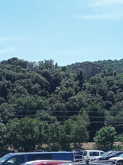 A stone house with a tiled roof sits atop a densely forested hill under a clear blue sky. The house has multiple windows, some rectangular and some angled. Below the hill, a line of trees is visible, and further in the foreground, several cars are parked. The environment appears to be a natural, hilly landscape with a residential structure integrated into it. The scene is likely in or around Besalú, Spain, given the architectural style and vegetation.