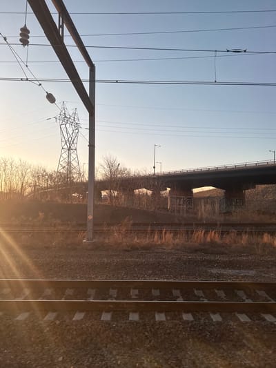 The image depicts an outdoor scene featuring railway infrastructure, a highway overpass, and electrical transmission lines, consistent with an urban-industrial edge environment in Philadelphia, Pennsylvania, USA, specifically along the I-95 corridor near the Schuylkill River.

In the foreground, two parallel electrified railroad tracks with concrete ties and dark ballast are present. Bright sunlight originating from the lower left creates a distinct lens flare effect on the tracks and ground.

The midground features an upward-sloping embankment covered in sparse, dry, brown vegetation, including bare deciduous trees and shrubs. A tall, gray metal pole with a small orange diamond-shaped marker is vertically positioned to the right of the tracks, supporting a complex overhead catenary system of wires and insulators that extends horizontally across the frame. A large, lattice-style electrical transmission tower is visible behind the bare trees on the left side of the embankment.

The background is dominated by a multi-lane concrete highway bridge supported by multiple concrete piers. Streetlights are visible along the bridge deck. Extensive, illegible graffiti covers the visible faces of the bridge piers, with discernible text including "I-95" and "SCH" on one pier. The sky transitions from a warm, golden-orange hue on the left, indicating a low sun angle (sunrise or sunset), to a clear, pale blue at the top right. No people or active vehicles are visible in the scene.