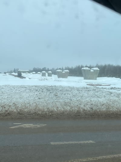 The scene depicts a winter landscape under an overcast sky, observed from within a vehicle, possibly in the city of Borisovo, Russia. The foreground shows a dark, wet asphalt road with a partial white line and a faded yellow marking, resembling a "7" or "Z," on its surface. Immediately adjacent to the road is a high, dirty snow bank composed of packed, brown-tinged snow.
Beyond the snow bank, the midground is dominated by a snow-covered, open construction site. Scattered across this terrain are multiple large, light grey concrete structures, identified as potential bridge supports or pillars. These structures vary; some are individual, wide-based pillars tapering upwards with concave, saddle-shaped tops designed to receive horizontal beams. Others appear as pairs of these structures. To the far left, a long, low, light-colored building is visible partially obscured by snow, alongside what appears to be a yellow piece of heavy construction equipment. Several reddish-brown objects, resembling timber or pipes, are lying on the snow to the right.
The background consists of a dense line of leafless trees forming a dark silhouette against the uniformly light grey sky, suggesting a wooded area or forest. No people are visible, and no active construction work or interactions are apparent. The environment is characterized by heavy snow cover and cold, diffuse lighting conditions typical of winter.