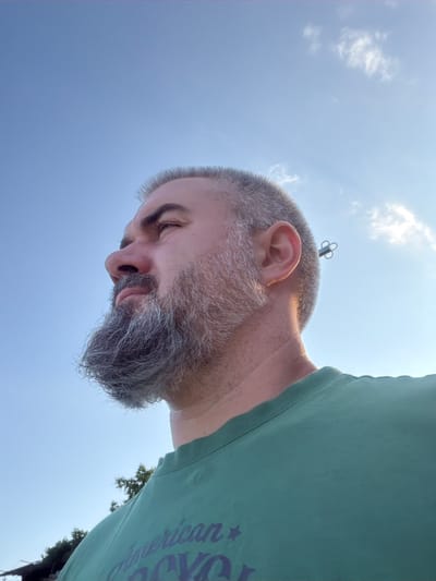 The image is an outdoor, low-angle, wide shot of a man with a beard and short hair. He is wearing a green t-shirt. The man is looking downwards and to his left, with his eyes closed. His face is in profile, and his beard is graying. A small, metallic object with a clover-like shape is visible on top of his head, possibly a hair accessory. The background is a clear blue sky with some wispy white clouds. The sun appears to be high in the sky, suggesting it is daytime. There are some green trees visible in the lower right corner of the image. The t-shirt has some text on it, which appears to be in purple lettering, and reads "American V." The overall mood of the image is relaxed and peaceful. The location is Gniazdovo, Bulgaria.