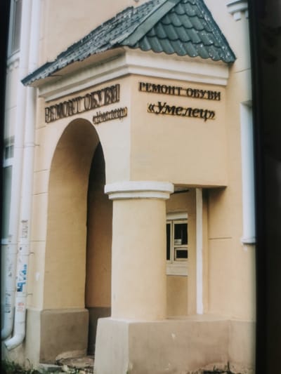 The image shows the exterior of a building with an arched entranceway. Above the entrance, a sign with gold lettering reads "РЕМОНТ ОБУВИ" which translates to "SHOE REPAIR." Below this, another sign in quotes says "«Умелец»" meaning "Craftsman" or "Handyman." The building has a light yellow facade and a dark tiled roof. A white column stands to the right of the entrance. Downspouts are visible on the left side of the building, one of which has some stickers on it. The scene appears to be in daylight, and the weather is not discernible. The image captures the storefront of a shoe repair business in Serov, Russia.