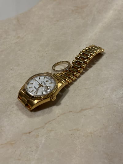 A gold-toned Rolex Oyster Perpetual Day-Date wrist watch, featuring a white dial, gold baton hour markers, a fluted bezel, and a Presidential bracelet, rests on a light beige marble-patterned surface. A gold-toned eternity ring, adorned with a continuous band of small, clear stones, is positioned atop the watch's bracelet near the watch case. The scene is located in the city of Tongxiang, China.