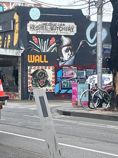 The image depicts a street scene in Balaclava, Australia, featuring a building with extensive graffiti art. On the building's dark facade, a sign reads "WEISHTZ KOSHER BUTCHERY SMALLGOODS MFR'S". Below this sign, there is artwork of red flowers and a black panther's head. To the right, a stylized depiction of a person spraying paint is visible. Further to the right, a utility pole stands, with a sign that includes the text "BALACLAVA KINDER". In the foreground, tram tracks run across a paved street, and a white traffic barrier is positioned in the lower portion of the image. A parked car, a bicycle rack with bicycles, and a utility box are also present. A person is visible through the window of a shop on the left side of the image. The overall environment suggests an urban setting.
