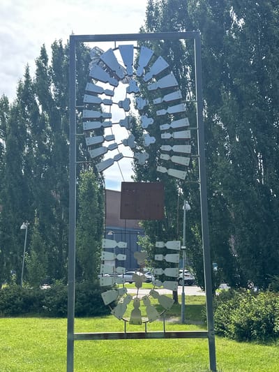 This image shows an outdoor sculpture on a sunny day in Hyvinkää, Finland. The sculpture is a large, abstract metal structure with numerous flat, angular pieces arranged in a circular, fan-like pattern. These metal pieces are a light, reflective color, possibly aluminum or stainless steel, and are attached to a dark grey metal frame. The sculpture appears to be kinetic, with the individual pieces designed to move in the wind.

In the background, there are tall, slender trees with dense green foliage, characteristic of poplars or similar species. Behind the trees, some buildings and parked vehicles are visible, suggesting an urban or suburban setting. The ground in front of the sculpture is covered in bright green grass, and there is also some landscaping with bushes and plants. A lamppost is visible to the left of the sculpture.

A small, weathered metal plaque is attached to the sculpture, though the text is difficult to read clearly. Some of the visible text appears to be "TH. JAKOBSEN" and "MINNES-GÅVA 1987". This indicates the sculpture is a memorial gift from Th. Jakobsen, dedicated in 1987.

The overall scene is bright and appears to be taken during daylight hours, with some clouds in the sky. The sculpture is the primary subject, dominating the center of the frame.