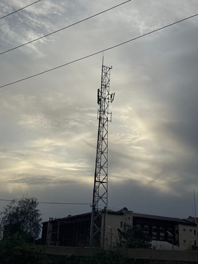 A low-angle, overcast shot shows a horizontal lattice boom crane pointing left across the frame. Thin, dark power lines cross diagonally in the foreground on the left. In the background is a building with multiple floors on the right, partially obscured by vegetation. The sky is overcast with striated clouds. The location is Obinze, Nigeria.