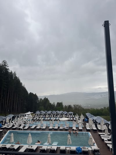 The image shows a view from above of an outdoor pool complex and surrounding landscape on an overcast day, likely in Sapareva Banya, Bulgaria. The foreground has a metal structure, with a row of pools running parallel to the right side of the frame. There are numerous white lounge chairs with umbrellas positioned around the pools, some of which are folded down. People can be seen in the pools. Beyond the pool area, there is a dense forest and a view of mountains obscured by mist or low clouds. The sky is predominantly gray and overcast.