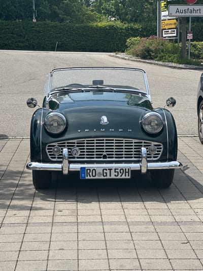 A dark green, convertible Triumph TR3 is parked on a paved area with a checkered pattern of light and dark grey square stones. The car is facing the camera, with its front bumper prominently displayed. The license plate reads "RO GT 59H" with a blue European Union band on the left and a "D" for Germany. Chrome accents are visible on the headlights, grille, and bumper. The car has a black soft top frame but the top is down. A single chrome antenna rises from the windshield frame. The background shows a grey asphalt road, a neatly trimmed green hedge, and trees. A signpost with multiple signs, including one that reads "Ausfahrt" (Exit), is visible to the right of the car. The lighting suggests a sunny day, casting shadows on the pavement.