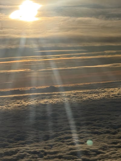 The scene is located over Hochheim, Germany, depicting an aerial view of a dense cloudscape. The sun, a bright, diffuse light source, occupies the upper left quadrant, emitting prominent light rays that descend diagonally towards the lower right. Multiple layers of clouds are visible: an upper section features elongated, horizontal strata with hues of gold and grey, indicative of high-altitude or low-angle sunlight. Beneath these, a vast, undulating blanket of densely packed, lower-level clouds extends across the lower two-thirds of the frame. The tops of these lower clouds are illuminated with golden light, contrasting with shadowed depressions. A distinct, circular green lens flare artifact is present in the lower right, superimposed over the cloud layer. No ground features, man-made structures, individuals, or aircraft are identifiable within the scene.