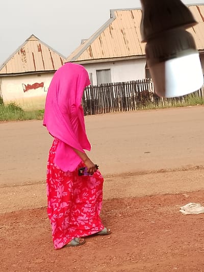 In this outdoor medium shot, a person wearing a vibrant pink hijab and a flowing red skirt with a floral pattern walks along a dirt and asphalt road in Agwala, Nigeria. The person is seen from the side, with their body angled slightly away from the camera. They are holding a purple object, possibly a phone, in their right hand. The person is wearing simple sandals.

In the background, two houses with corrugated metal roofs, exhibiting signs of rust and age, are visible. A rustic wooden fence separates the houses from the road. Graffiti, including what appears to be the letters "D" and "L", is present on the side of one of the houses. The scene suggests an overcast or hazy day, with the sky a pale, washed-out white. The ground beside the road is a mix of reddish-brown dirt and gravel, with some sparse green vegetation visible in the distance. A crumpled white piece of fabric lies on the ground to the right of the person. The overall impression is one of a casual, everyday moment in a rural or semi-urban Nigerian setting.
