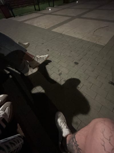 The image shows a low-angle view of several pairs of feet wearing white sneakers, with shadows cast on a paved area. The paving consists of rectangular grey stones arranged in a grid. In the background, a dark wooden bench and greenery are visible, suggesting an outdoor setting, possibly a park or a public seating area at dusk or night. The lighting creates distinct shadows of the people present, some of whom appear to be wearing jeans or patterned trousers. One individual's leg in the foreground exhibits visible veins and tattoo markings. There are no other discernible objects or activities in the immediate vicinity. The location of Siedlce, Poland, is not evident from the visual cues in the image.