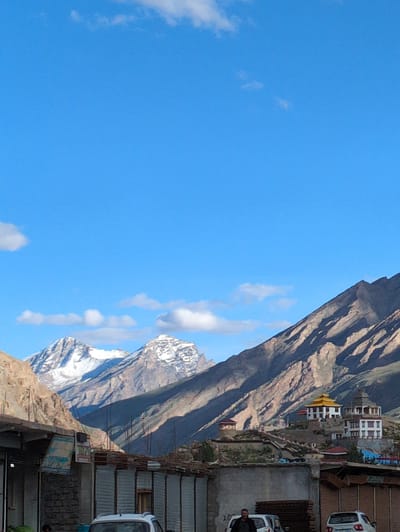 A vertical, eye-level shot captures the sprawling landscape and buildings in the city of Padum, India. The sky is a clear, bright blue with scattered white clouds. In the background, majestic, snow-capped mountains rise, their slopes casting long shadows under the sunlight. Nestled on the mountainside is a white monastery with a distinctive yellow roof and a stupa. In the foreground, the lower portion of the image shows a street scene with buildings made of corrugated metal and brick, some with faded signage. A white car is parked in front of one of the buildings, and a person can be seen standing near it. Another white car is visible further down the street. The overall impression is one of a remote and serene mountain town.