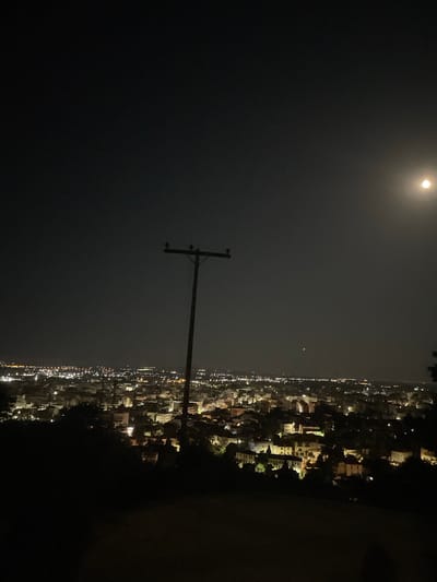 This is a nighttime, wide-angle shot of the city of Xanthi, Greece, taken from an elevated viewpoint. The foreground is dominated by a silhouetted hillside covered in dark foliage, obscuring any details of the terrain. Rising from the center of the frame is a prominent wooden utility pole with insulators, starkly silhouetted against the dark sky.
In the midground, the city of Xanthi sprawls across the landscape, a sea of illuminated buildings and streetlights creating a warm glow against the dark surroundings. The architecture appears to be a mix of residential and commercial structures, with visible rooftops and window lights. The density of buildings suggests a vibrant urban area.
The sky is a deep, inky black, characteristic of a clear night. A bright, full moon hangs in the upper right quadrant of the image, casting a soft, ethereal light over the scene. The moon's luminosity creates a subtle haze in the sky around it, and a very faint, distant light appears to the left of the moon, possibly a planet or a very distant aircraft. The overall impression is one of a peaceful, illuminated cityscape under a celestial glow.