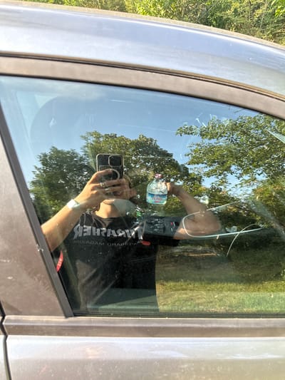 In Rudiano, Italy, a person is reflected in a car window, holding a smartphone and a bottle of "Flavia" brand water. The individual is wearing a black t-shirt with white lettering that reads "THRASHER" in reverse. They have a watch on their left wrist and several rings on their fingers. The car's interior is visible in the reflection, including a portion of the dashboard. Outside the car, trees and a bright blue sky are visible. The car appears to be parked in an outdoor setting, possibly a parking lot or roadside, given the presence of greenery and natural light. The image is taken from a low angle, focusing on the reflection in the window.