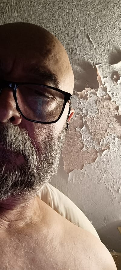 The image shows a close-up, low-angle, selfie-style shot of a bald, middle-aged man with a graying beard and glasses. He is facing the camera, with his left eye and part of his nose visible. The background is a textured, peeling wall, suggestive of an older building. The lighting is dim and directional, casting shadows across the man's face and highlighting the texture of the wall. There is a hint of a light-colored pillow or bedding on the right side of the frame, suggesting the man is lying down or reclining. The scene appears to be indoors, with no visible windows or outdoor elements. The location is described as Vironas, Greece, suggesting the photograph was taken within a residential or public building in that area. No specific landmarks or distinguishing features of Vironas are evident in the image itself.