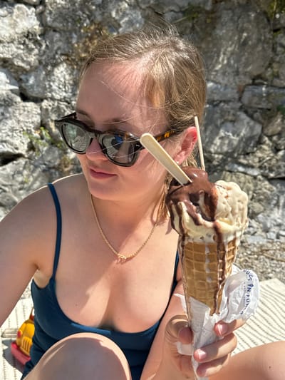 A female subject, appearing to be an adult with light brown hair, is depicted in a close-up outdoor scene in Abbadia Lariana, Italy. She wears tortoise-shell sunglasses and a dark teal ribbed top, accessorized with a delicate gold-colored chain necklace featuring a small central element. Her gaze is directed to her left, with a relaxed expression.
The subject's right hand holds a large ice cream cone. The ice cream consists of multiple scoops in at least two flavors (one light, one dark), heavily drizzled with dark chocolate sauce that is visibly melting and dripping down the sides of the scoops and waffle cone. Two small, light-colored wooden spades are inserted into the ice cream. The lower portion of the cone is wrapped in white paper, displaying partial text including "S IN FU" and "GASTRONOMIA."
The background features a sun-lit, rough-hewn stone wall or rock face. The ground in the foreground is covered by a light-colored, striped fabric, possibly a blanket or towel. A yellow and red plastic toy is partially visible on the ground to the lower left. The overall lighting is bright, consistent with direct sunlight on a clear day, indicating a warm environment. The scene is consistent with a recreational area, likely near the shores of Lake Como in Abbadia Lariana.