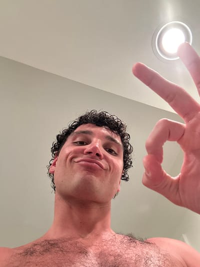 The image shows a man with curly brown hair, making an "ok" sign with his right hand towards the camera. He is wearing no shirt and has a light brown complexion with light hair on his chest. The man's facial expression suggests he is smiling slightly. The background is a pale green wall with a white ceiling. There is a bright, white, circular light fixture in the ceiling to the right. The image appears to be a selfie taken in a bathroom or a room with a bathroom-like appearance, at 25.773 latitude and -80.14 longitude, located in Miami Beach, Florida, United States.