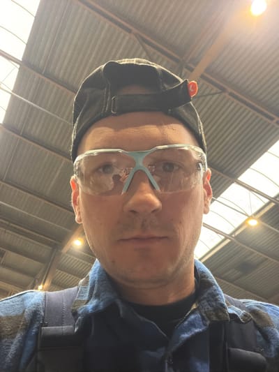 A man wearing safety goggles and a black cap is looking directly at the camera. He is wearing a blue shirt or jacket. The background consists of a high, corrugated metal ceiling with skylights, suggesting an industrial building. Given the request to place the image in Bydgoszcz, Poland, it is plausible this photo was taken inside a factory, warehouse, or large construction site within the city.
