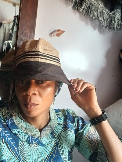 This image depicts a person, appearing female, in an indoor setting in Sabon Gari South, Nigeria. The subject, possessing a dark skin tone, is centered in the frame, looking directly at the camera with a neutral expression. They wear a two-tone bucket hat; the upper section is light brown with three horizontal black stripes, while the brim is dark brown fabric. The subject's left hand is raised, with fingers gripping the brim of the hat, suggesting an adjustment. They are dressed in a patterned garment, likely a tunic, featuring shades of blue and light green with a textured, light-colored collared neckline. Silver-colored hoop earrings are visible, and a white earbud is inserted into the right ear. A black wristwatch with a dark rectangular face is worn on the left wrist. The background on the left side shows a vertical wooden frame, appearing to be a mirror frame, reflecting a piece of hanging clothing. On the right, a plain white wall is visible, displaying a brown rectangular tag, possibly labeled "SENO," and a draped black and white fringed textile in the upper right corner.