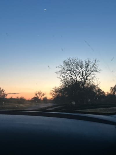 The image depicts a landscape scene with a crescent moon visible in the upper left corner of a light blue sky. A tree with bare branches is silhouetted against the sky. The sky transitions into a gradient of orange and yellow towards the horizon. Additional trees and vegetation are visible along the horizon line. The right side of the image is obscured by what appears to be part of a vehicle interior, with a dark dashboard or surface. The location is not identifiable as Krasnovo, Bulgaria from the image. No people, subjects, objects, actions, or interactions are discernible in the image beyond the landscape.