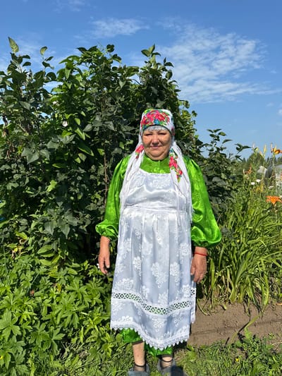 An elderly woman stands in a garden in Sredneye Kechevo, Russia, on a sunny day. She is wearing a bright green dress, a white apron with a floral pattern, and a colorful headscarf. The garden is lush with green foliage and flowers, including orange lilies. The sky is blue with scattered white clouds. The woman is looking directly at the camera with a slight smile. The overall scene is peaceful and idyllic.