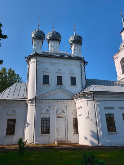 The image shows the exterior of the Church of the Nativity of the Most Holy Theotokos in Kady, Russia. The white stone church has multiple domes with crosses on top, and a grey sloped roof. The church features several windows with brown frames and intricate patterns, as well as a white door with a small set of steps leading up to it. The building is surrounded by green grass and a few trees are visible in the background. The sky is clear and blue.