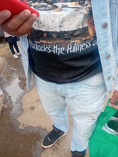 The image presents a close-up view of a person standing on a wet, paved surface in Nanyuki, Kenya. The individual is wearing a light blue denim jacket over a black t-shirt. The t-shirt features a graphic print: an upper portion depicting various objects on a white background, and a lower portion with white text on a black background, which reads "unlocks the fullness of". Light blue denim jeans, exhibiting some light distressing on the left thigh, cover the legs. Black athletic shoes with white soles and red accents are worn on the feet. The person's left hand holds a red rectangular object. The right hand grasps a bright green reusable bag, which contains a reflective item. In the blurred background to the left, the lower legs and feet of a second individual, clad in dark trousers or a skirt and white sneakers, are partially visible. The ground beneath is concrete, characterized by extensive wet patches and reflective areas, indicating recent moisture. Several small, green, indistinct objects are visible on the wet surface.