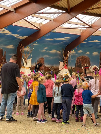 This image depicts an indoor event space decorated with a Western or Native American theme. The ceiling features exposed wooden beams and translucent panels, with colorful string lights hanging down. The backdrop is a mural of a desert landscape with mesas and a blue sky with clouds.

In the foreground, a group of children and a few adults are gathered on a sandy floor strewn with confetti. Many of the children are wearing headdresses and sashes, suggesting a themed costume party or performance. Some of the children have face paint, resembling war paint.

Several teepee structures are visible throughout the space, some appearing to be decorative props and others potentially part of the activity. One teepee in the background is adorned with colorful ribbons. A man in a feathered headdress and traditional attire is visible in the middle ground, facing away from the camera.

The overall atmosphere appears festive and engaging for children, likely part of an organized event. The location is identified as the Speelstad Oranje theme park in Collendoorn, Netherlands, which is known for its indoor play areas and family-friendly events.