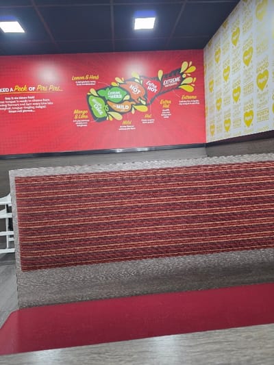 The image depicts the interior of a Pepe's Piri Piri restaurant, specifically located at 30 Bore St, Lichfield WS13 6LZ, United Kingdom. In the foreground, a red upholstered booth seat is visible. Directly behind it, a booth backrest features horizontal stripes in shades of red and brown, mounted on a grey wood-effect laminate base. The floor is also covered in grey wood-effect laminate. The left wall displays a large red mural detailed with text promoting Piri Piri flavors and a diagram illustrating spice levels ranging from "Lemon & Herb" and "Mango & Lime" through "Mild," "Hot," "Extra Hot," to "EXTREME INFERNO." The right wall is adorned with white wallpaper patterned with repeating yellow heart shapes containing "PEPE'S Good Worth The Wait!" branding. The ceiling is dark, comprising black tiles with multiple recessed square LED light panels. No individuals or specific actions are present in the scene.
