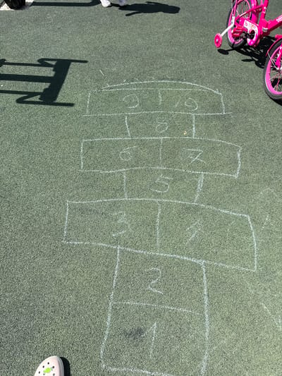 The image shows a hopscotch grid drawn with white chalk on a green, artificial turf surface. The grid consists of numbered squares and rectangles, with numbers visible including 1, 2, 3, 4, 5, 6, 7, and 9. In the background, there are shadows cast by playground equipment and two pink children's bicycles with training wheels. The scene appears to be an outdoor recreational area, likely a playground or park. The specific location in the city of Вязники, Russia is not identifiable from the image.