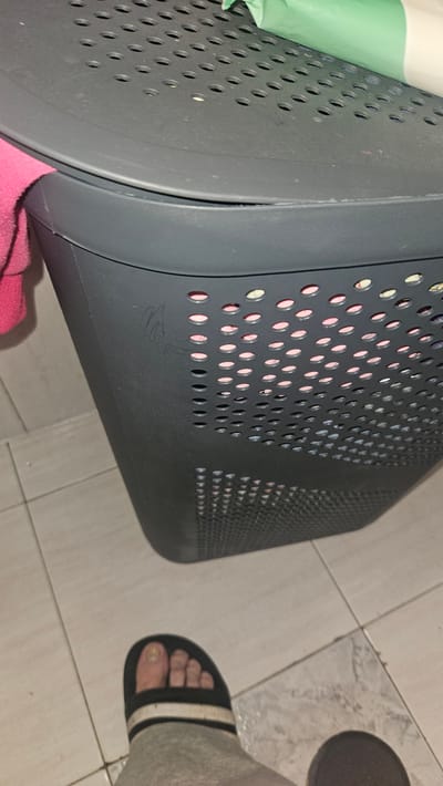A close-up, low-angle shot focuses on a dark gray laundry basket with a perforated lid, set against a tiled floor. A pink towel is draped over the left side of the basket. The basket's surface has a faint, scrawled marking. Inside the basket, visible through the perforations, are items of clothing in various colors, including pink and green. In the foreground, a person's foot wearing a black and white striped sandal is visible, resting on the tiled floor. The scene is indoors, likely a bathroom or laundry room, given the tiling. The image was captured in Kiryat Yam, Israel.