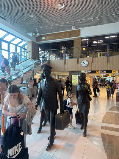 The image depicts the interior of an airport terminal in Bubuieci, Moldova. In the foreground, there are bronze statues of a man and a woman, seemingly travelers with luggage. The man, dressed in a suit and hat, holds a suitcase. The woman, in a skirt suit, also holds a suitcase. Several living people are present in the scene, including women and children. One woman, in a white top, is seen with a black Nike backpack. Another young girl, in a white t-shirt and shorts, stands near the statues. In the background, there are escalators, signage indicating "Ascensor Elevator" and "Sosiri Arrivals", and a large clock on the wall. People can be seen walking and standing throughout the terminal. The overall environment suggests a busy airport setting with modern architecture and flooring.